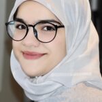 Photo depicts a woman with black glasses, smiling at camera wearing a white headscarf and light top.
