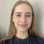 Image depicts a woman smiling at the camera. She has long blonde hair, a dark blue top, and is standing in front of a white wall.