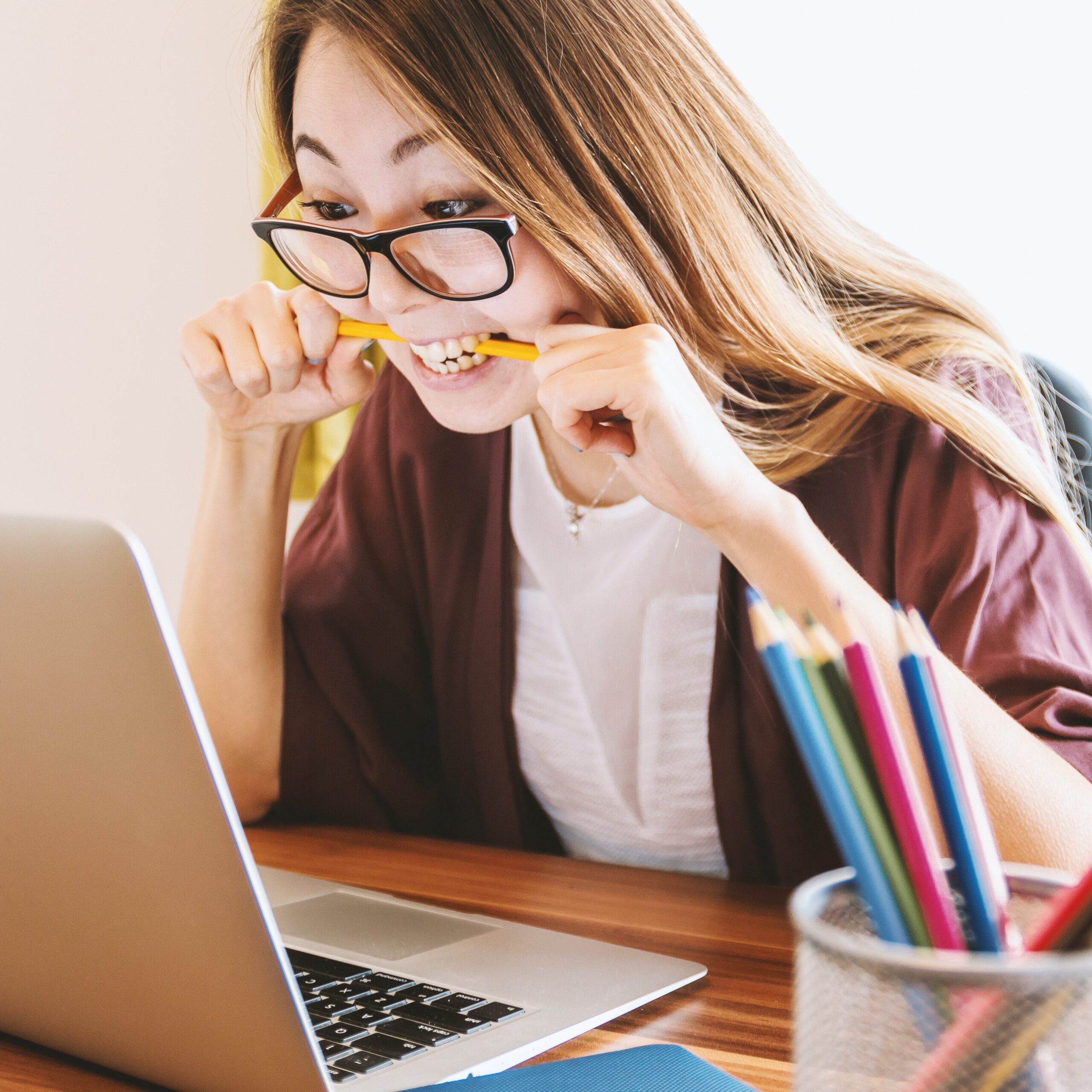 Photo depicts a female-presenting Asian person with black glasses and long light hair staring at their laptop screen biting a pencil looking frustrated.