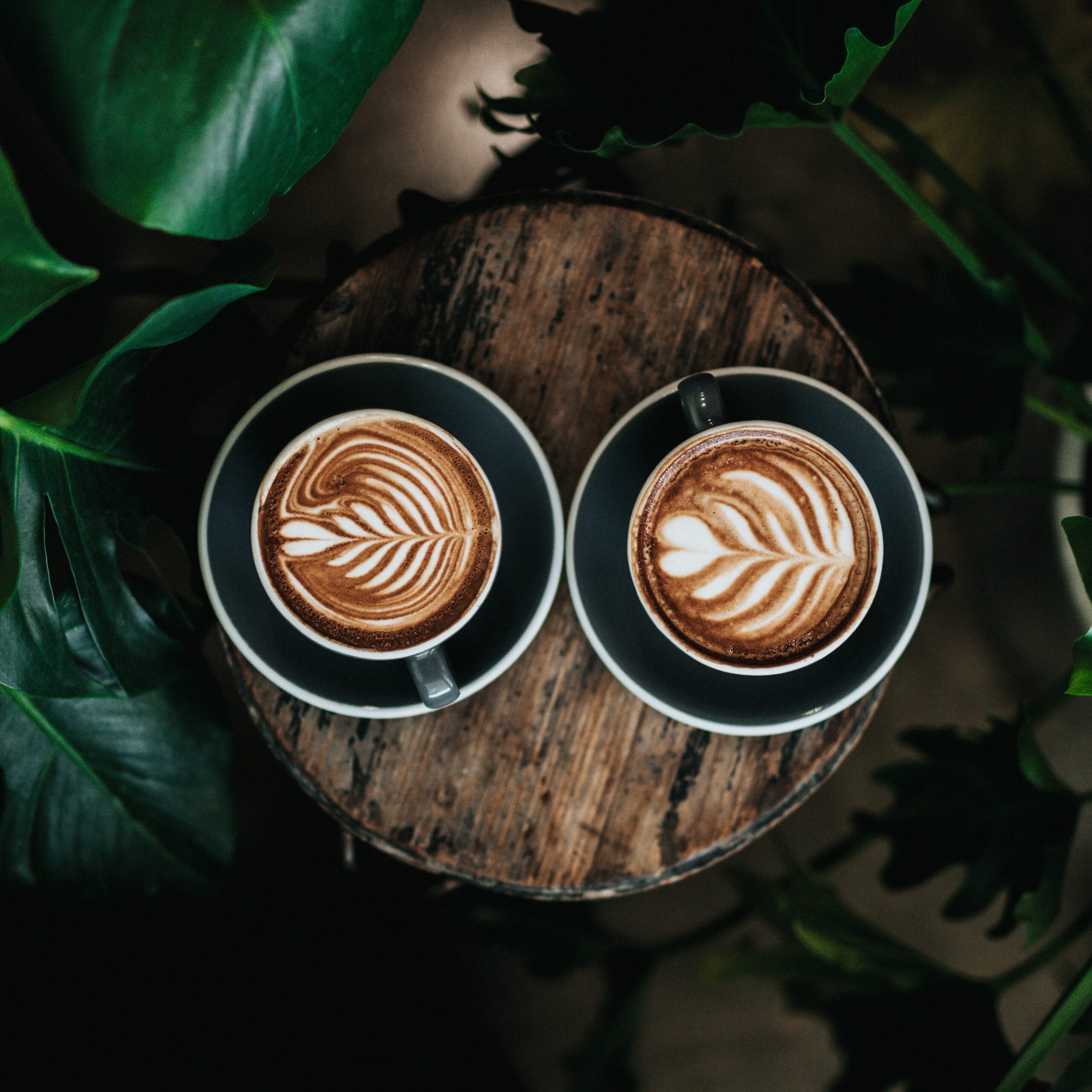 Top-down photo of two coffee mugs on saucers, placed on a worn wood block surrounded by leaves. The coffee has a pattern made in the foam.