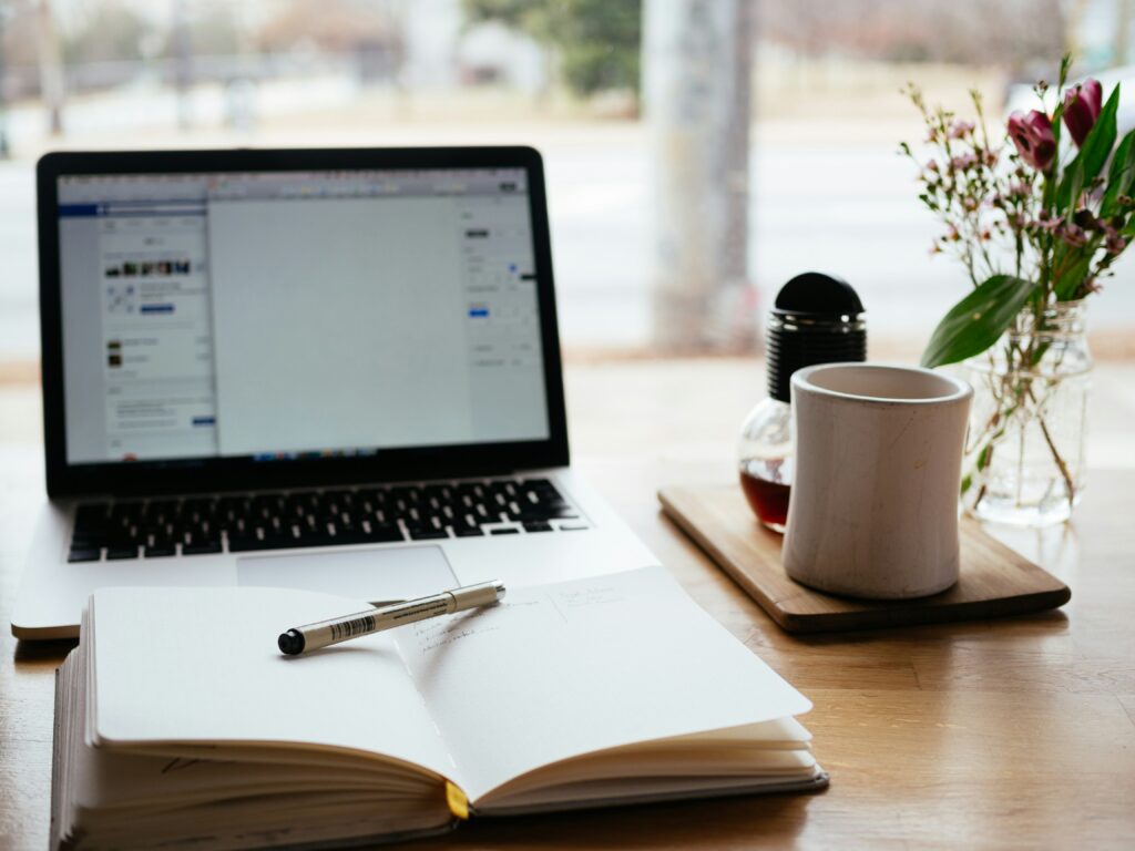 A laptop with a notebook and pen set aside from a coffee mug. 