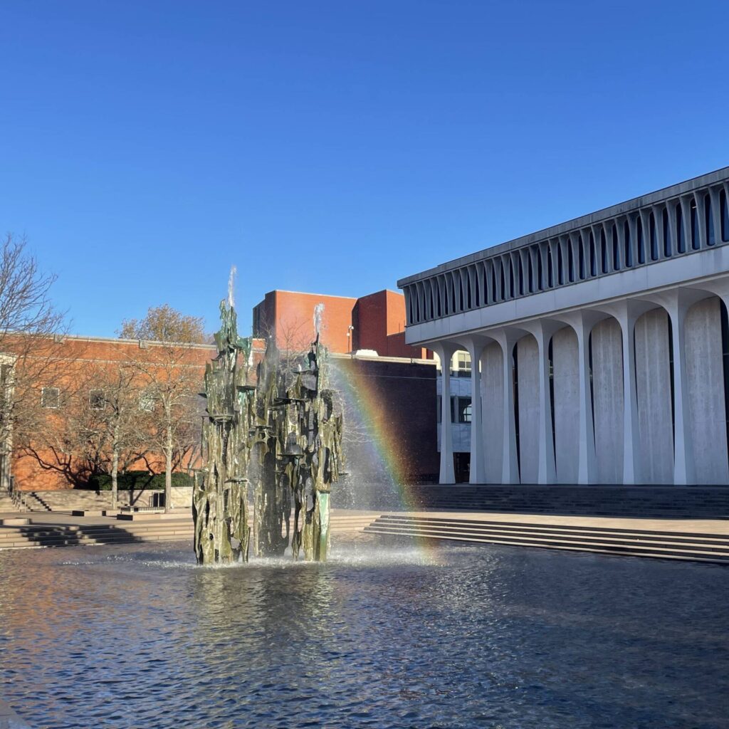 Image of a fountain in the foreground whose spray creates a rainbow. Background includes the School of Public and International Affairs